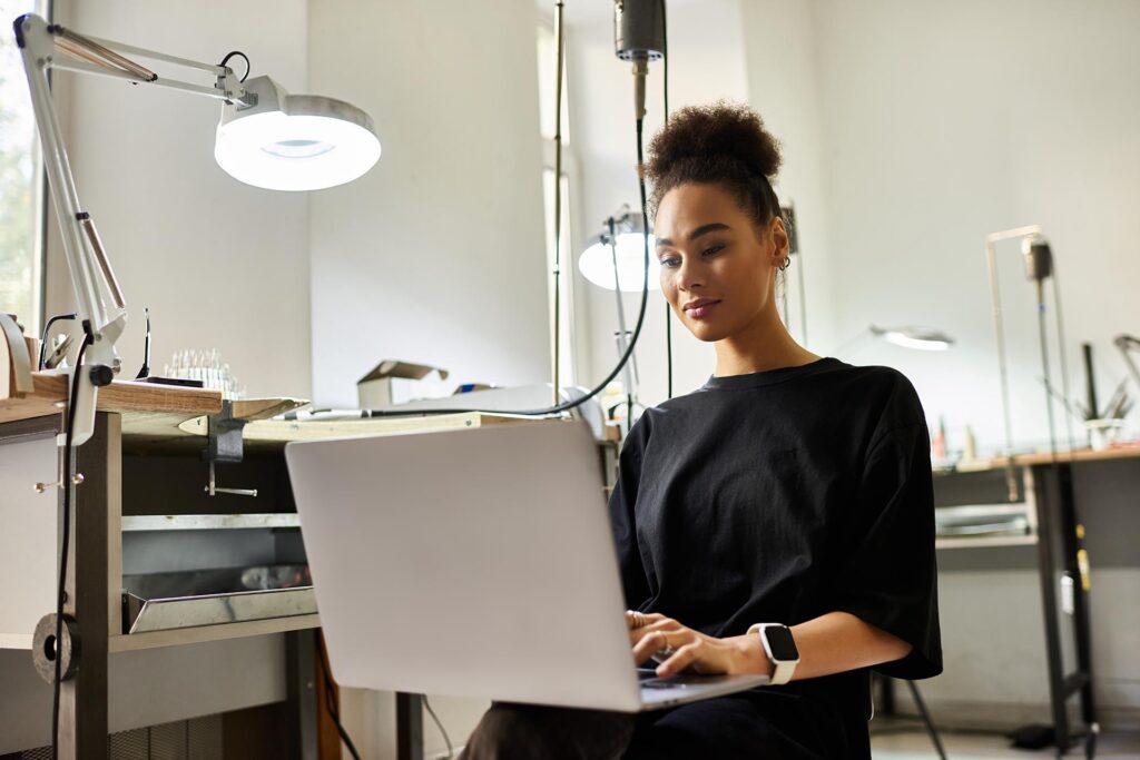 A talented woman types on her laptop in a jewelry workshop, showcasing creativity and dedication.